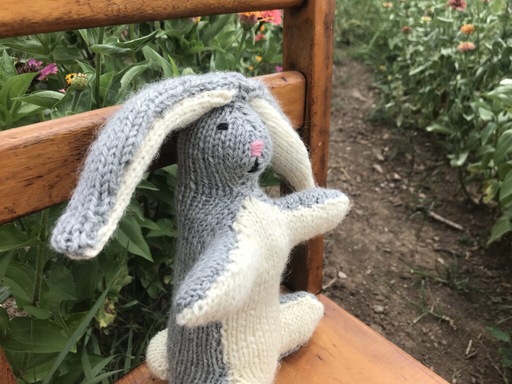 grey and white knitted rabbit sitting in antique child's chair in a flower garden