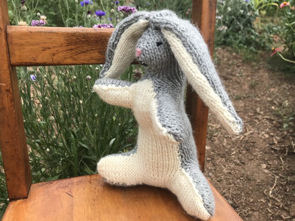 grey and white knit rabbit facing left on brown wooden chair in flower garden