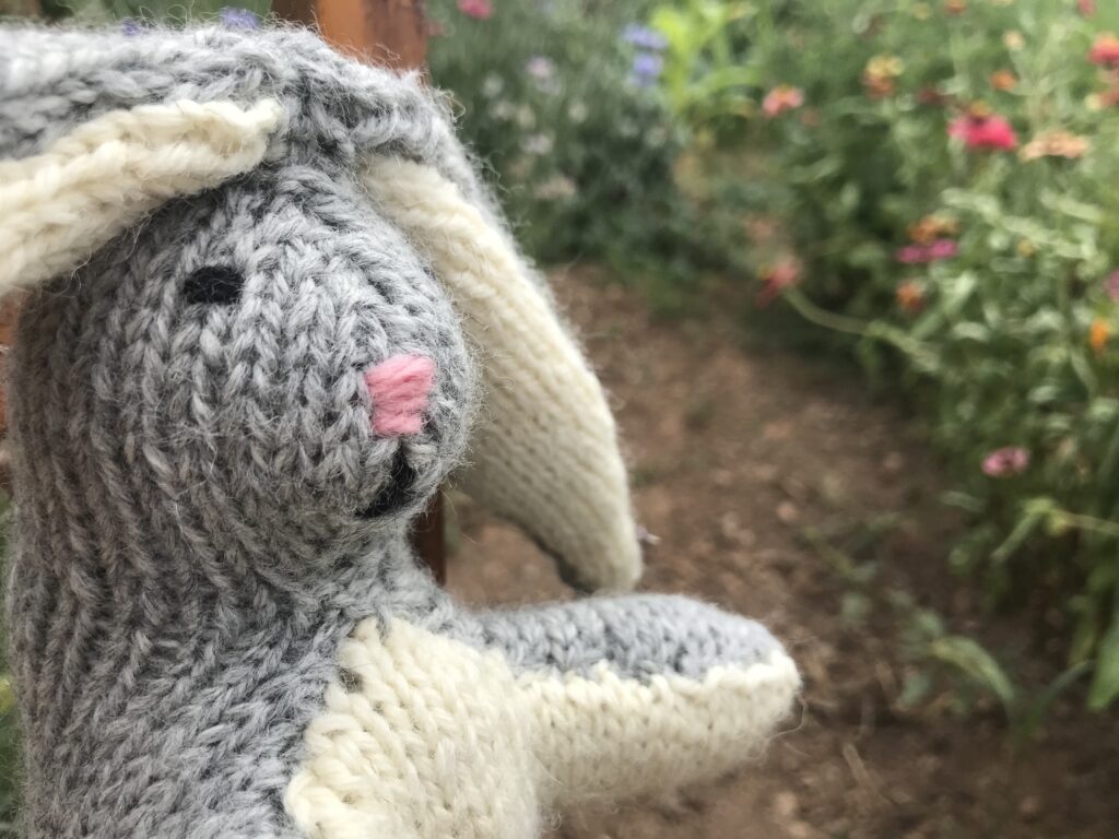 closeup of grey and white knit rabbit on brown wooden chair in flower garden