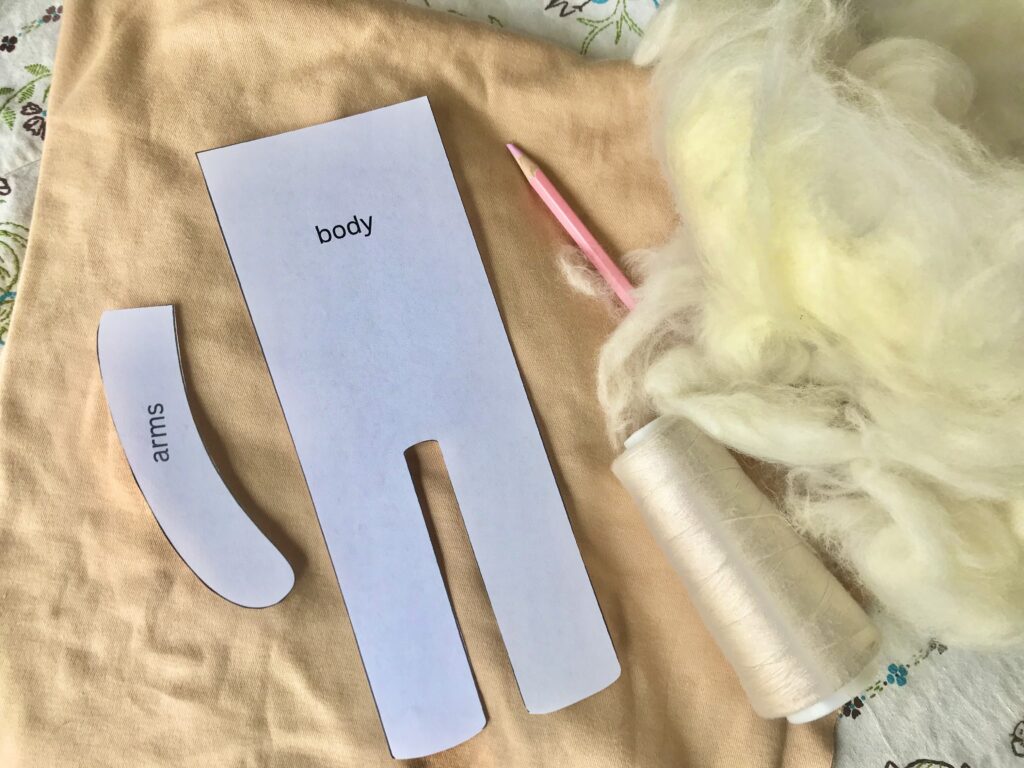 supplies needed to make a waldorf doll body including wool, cotton interlock fabric, waldorf doll body pattern, thread, chalk pencil, laid out on an embroidered tablecloth