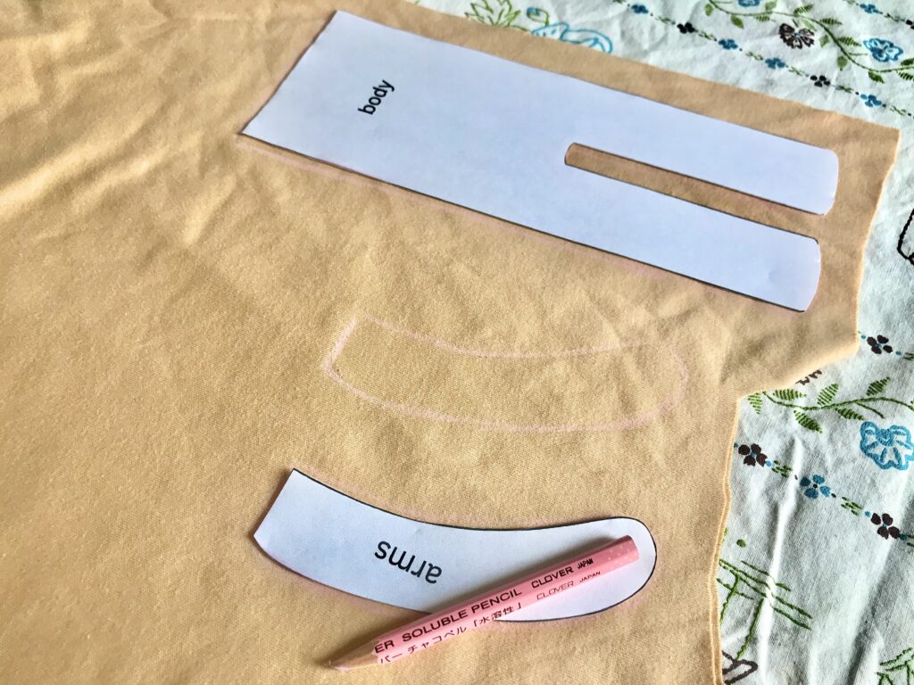 doll body and arm pattern pieces laid out onto fabric being traced with a pink pencil on a tablecloth