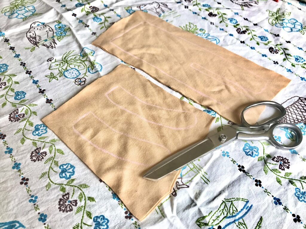 traced doll pieces on fabric with scissors on a blue and green and white embroidered tablecloth