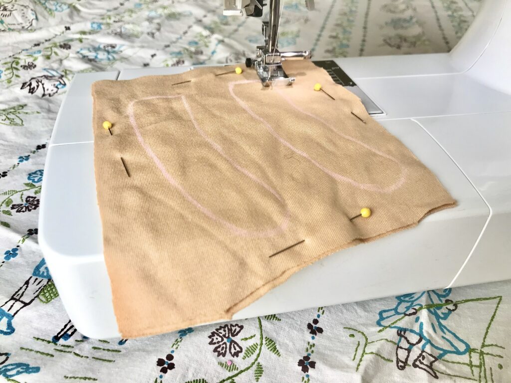 tan fabric with pink pencil markings being sewed with an electric sewing machine with yellow pins and a multicolored tablecloth