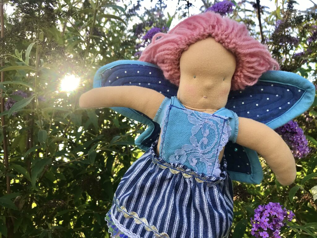 Waldorf Butterfly fairy doll pink hair blue dress blue wings with butterfly bush and sunshine in the background