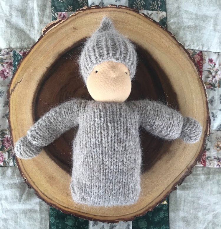 grey knitted waldorf doll laying on a sliver of log and quilt