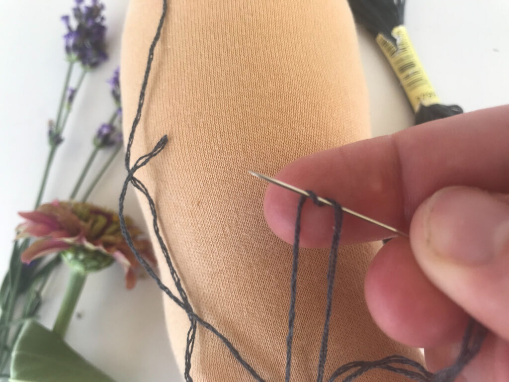 woman's hand tying a french knot with unfinished waldorf doll head and fresh flowers in background