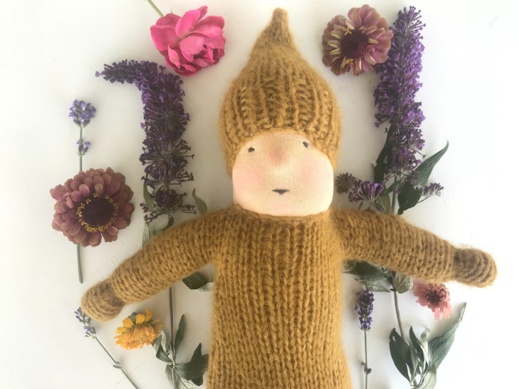 Mustard Yellow knitted waldorf bunting doll wearing hat with purple and pink flowers in the background