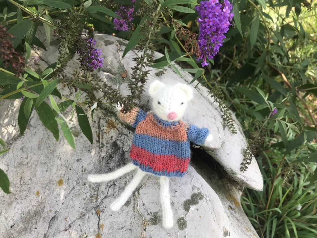 Knitted mouse wearing sweater on rock with butterfly bush in the background