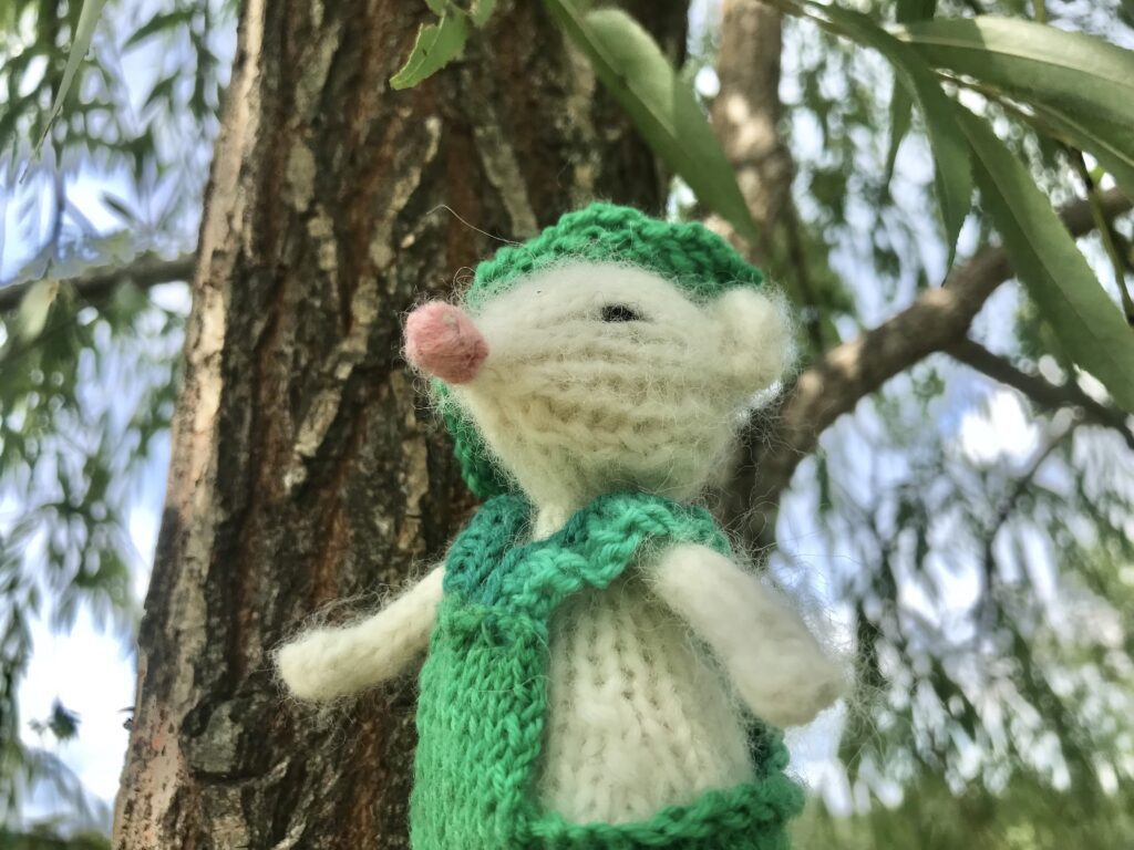 white knitted mouse in green knitted striped overalls sitting in a tree