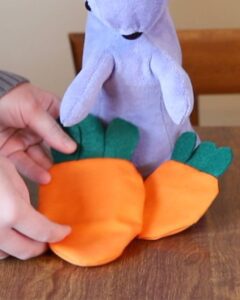 woman's hand laying orange fabric fillable easter egg on a brown wood table on top of a purple rabbit toy
